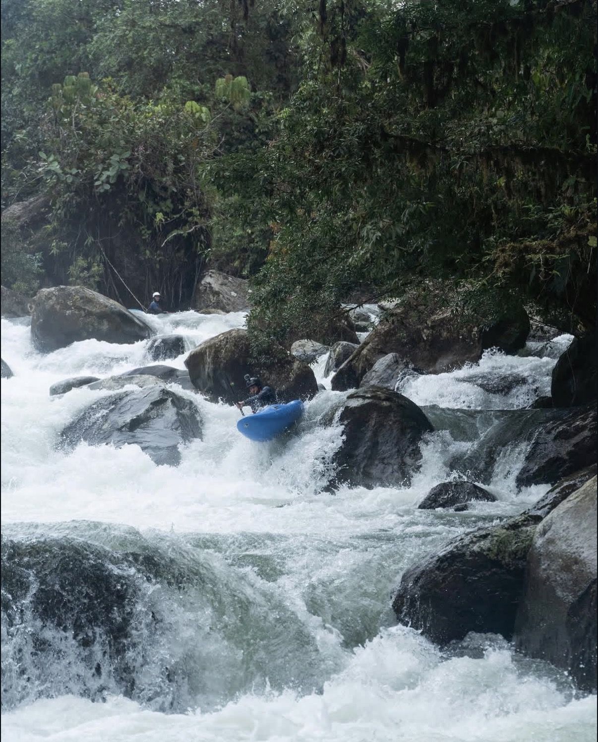 A kayaker in a blue boat threads a steep boulder rapid as a safety paddler watches