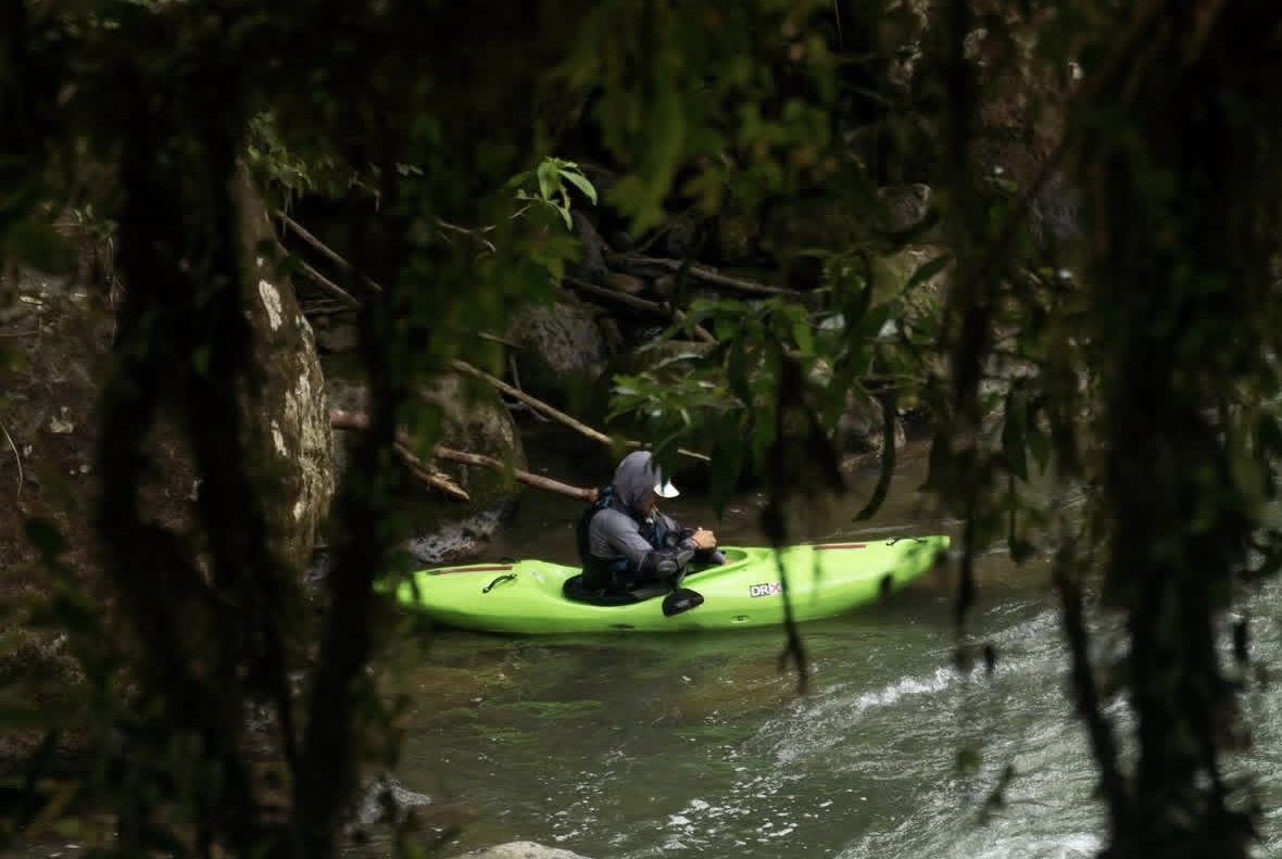 A kayaker in a green boat waits in an eddy, seen through the forest